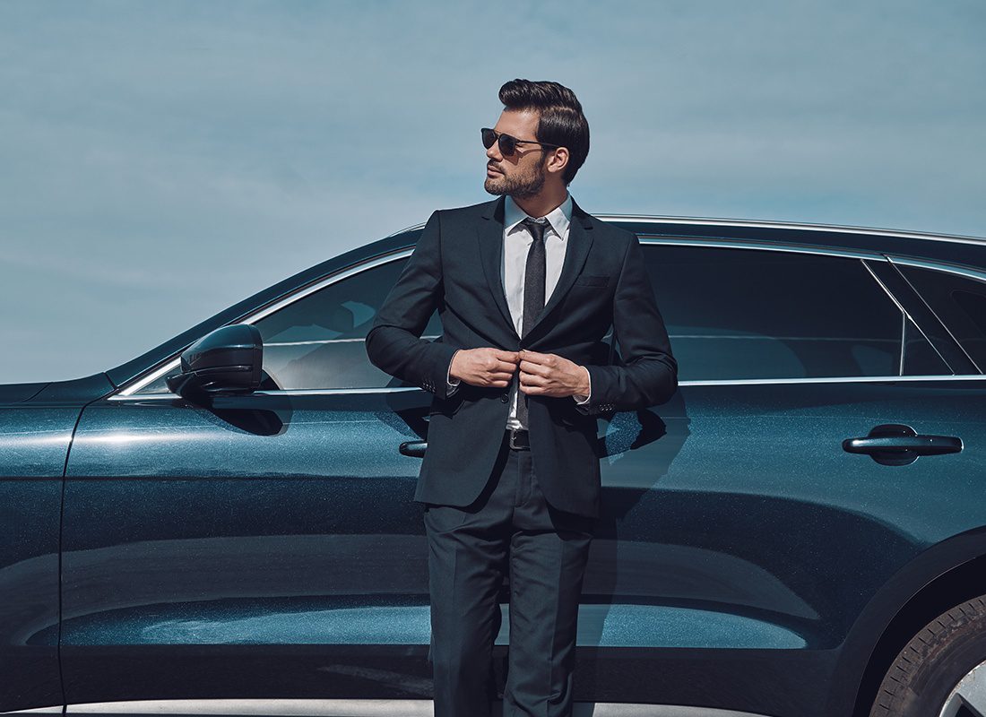 Business Insurance - Man Wearing a Black Suit and Sunglasses Leans on a Black High-End Car on a Sunny Day